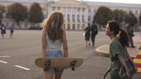 portrait of two girls Teenage friends with skateboard in sunshine in street european town laughing at something outside and talk. slow motion