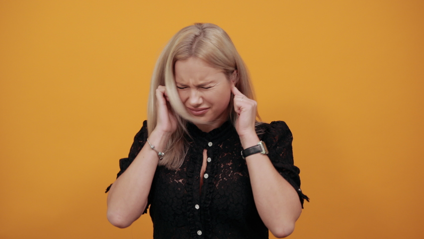 Beautiful blonde girl in black dress on yellow background irritated angry woman covered ears with fingers to avoid hearing sound of noise