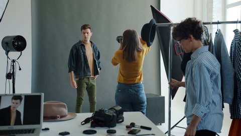 Backstage of studio photoshoot: handsome guy model is posing for male photographer while stylist is moving clothing on hanger. People and advertising concept.
