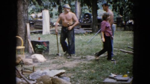 WAUCONDA ILLINOIS USA-1960: Two Shirtless Men Trade A Sledge Hammer For A Rifle While Working In A Yard