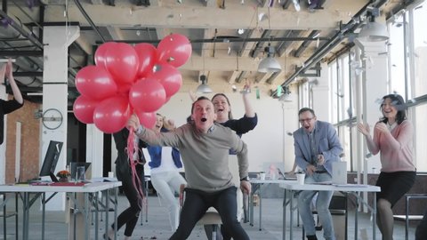 Colleagues congratulate the young manager. They give him red balloons, ride them in an office chair, and blow up crackers with confetti. Business team dancing and celebrating. Corporate. Office life