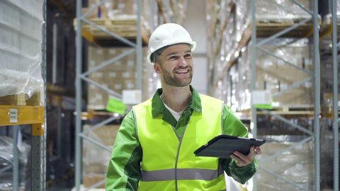 man warehouse worker walks through rows of storage racks with merchandise and looks at digital tablet. male smiling. architect walking through logistics shelves of factory. male in storage with goods