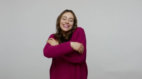 Self-care and love. Attractive charming brunette woman in pullover embracing herself, dreaming pleasant memories, smiling with satisfied expression. indoor studio shot isolated on gray background