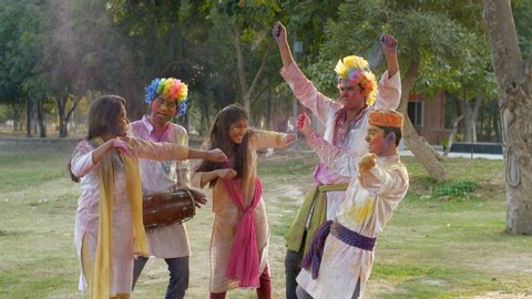 Cheerful young friends enjoying Holi festival celebrated in India - festive scene. Group of funny Indian teenagers smeared in Gulal colors, happily dancing together and playing Dholak during a Holi...