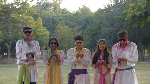 Cheerful Indian teenagers happily playing with colorful Gulal during Holi celebration. Group of young friends in cool sunglasses blows Holi powder colors off their hands and wishes Happy Holi looki...