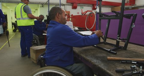 Front view of an African American male worker in a workshop at a factory making wheelchairs, sitting at a workbench assembling the frame of a product and checking it, he is sitting in a wheelchair,