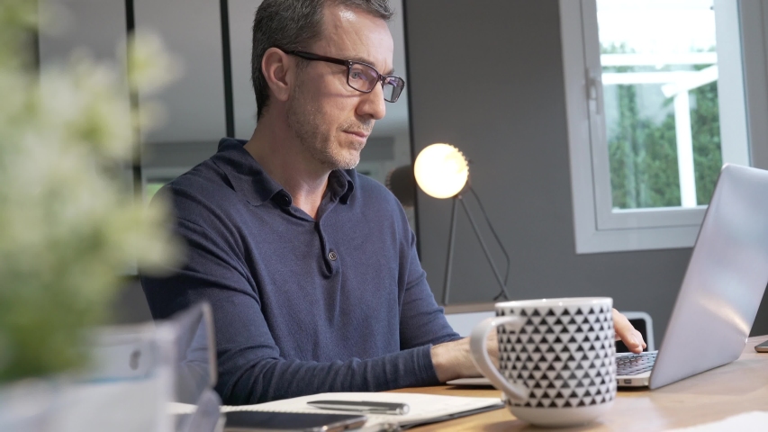 Middle-aged guy having hot drink, working on laptop Royalty-Free Stock Footage #1046320900