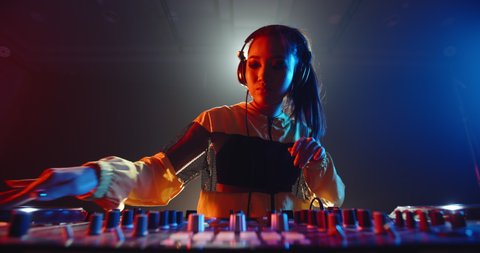 Cool asian female disc jockey standing at mixer cotroller, creating a music list in a nightclub, lit by red and blue lights - nightlife concept 4k footage