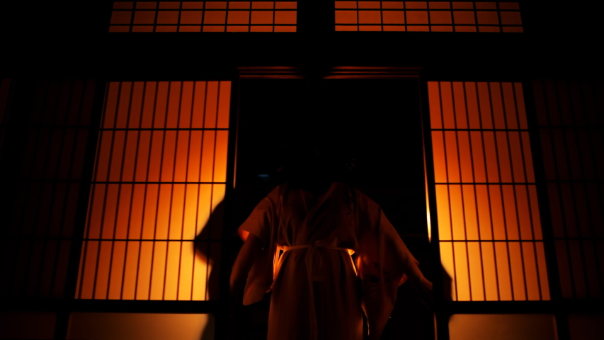 Geisha or maiko silhouette behind sliding paper doors with colorful backlighting opening door and entering room hall at night in wooden traditional Japanese house