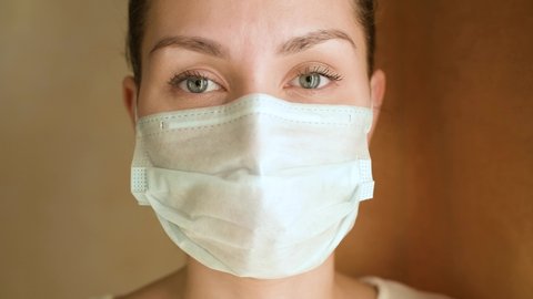 Close-up of a young woman in a protective medical mask, she looks directly at the camera and then turns to the window