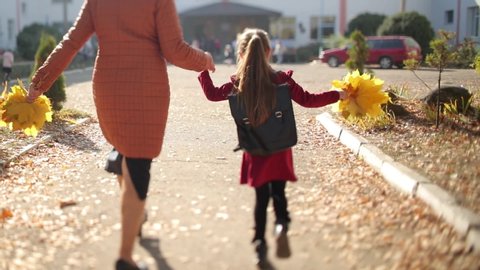 Стоковое видео: Mother and a little girl in red dress with a backpack on shoulders and a bouquet of yellow autumn leaves in hand go to school. Sunny autumn. 4K video.