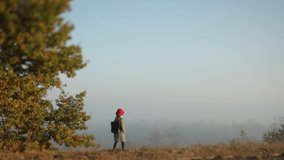 Foggy autumn scenery. Small pretty girl in grey coat and red hat walks slowly through autumn forest in sunny day. 4K video.