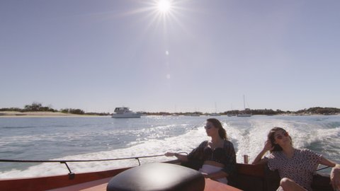 Two beautiful young women wearing sunglasses cruising on a motorboat near the beach, between other bigger boats, under a sunny blue sky. Medium to long shot on 4K RED camera with lens flare.
