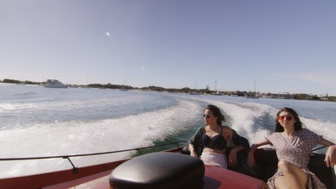 Two beautiful young women wearing sunglasses cruising on a motorboat near the beach under a sunny blue sky. Medium to long shot on 4K RED camera with lens flare.