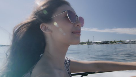 Beautiful young woman wearing sunglasses smiling, cruising on a motorboat, wind in her hair, near the beach under a sunny blue sky. Close shot on 4K RED camera with lens flare.