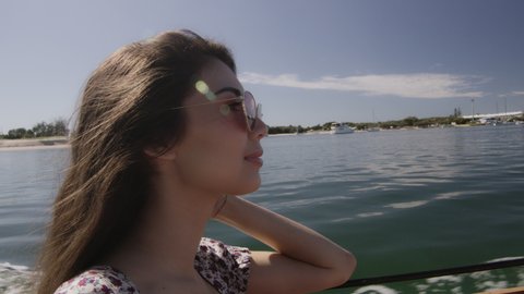 Beautiful young woman wearing sunglasses cruising on a motorboat, wind in her hair, near the beach under a sunny blue sky. Close to medium shot on 4K RED camera with lens flare.