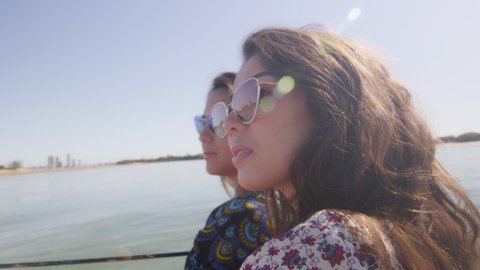 Two beautiful young women wearing sunglasses cruising on a motorboat near the beach under a sunny blue sky. Close shot on 4K RED camera with lens flare.