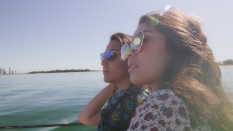 Two beautiful young women wearing sunglasses cruising on a motorboat near the beach under a sunny blue sky. Close shot on 4K RED camera with lens flare.