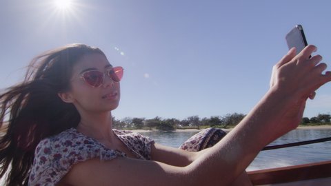 Beautiful young woman wearing sunglasses cruising on a motorboat, wind in her hair, taking selfies, near the beach under a sunny blue sky. Medium shot on 4K RED camera with lens flare.