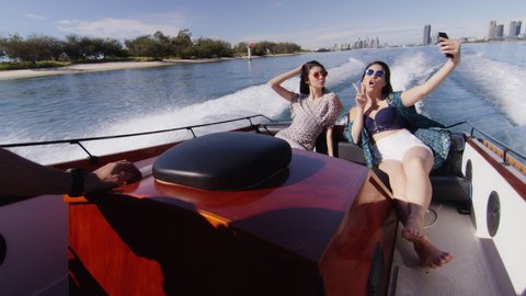 Handsome young man and two beautiful young women wearing sunglasses cruising on a motorboat, taking selfie near the beach under a sunny blue sky. Close to long shot on 4K RED camera with lens flare.