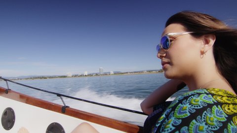 Beautiful young woman wearing sunglasses cruising on a motorboat, wind in her hair, passing the hand in her hair, near the beach under a sunny blue sky. Close to medium shot on 4K RED camera.