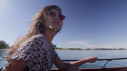 Beautiful young woman wearing sunglasses cruising on a motorboat, passing the hand in her hair, near the beach under a sunny blue sky. Close to medium shot on 4K RED camera with lens flare.