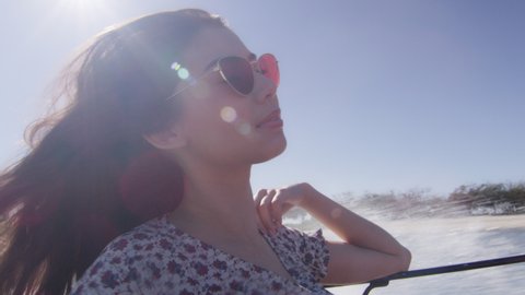 Beautiful young woman wearing sunglasses cruising on a motorboat, wind in her hair, near the beach under a sunny blue sky. Close to medium shot on 4K RED camera with lens flare.