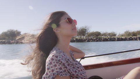 Beautiful young woman wearing sunglasses cruising on a motorboat, wind in her hair, near the beach under a sunny blue sky. Medium shot on 4K RED camera with lens flare.
