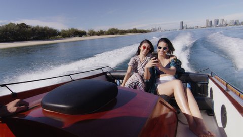 Handsome young man and two beautiful young women wearing sunglasses cruising on a motorboat, laughing and taking selfie near the beach under a sunny blue sky. 4K RED camera with lens flare.