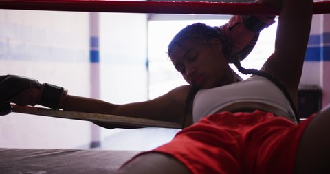 Front view close up of a mixed race female boxer with long plaited hair at a boxing gym, lying against the ropes in a boxing ring after being hit, backlit, slow motion