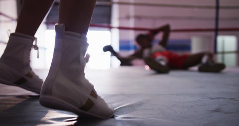 Side view low section of the boots of a female boxer moving and jumping in a boxing ring, her oponent lying on the ropes, knocked out on the other side of the ring, slow motion