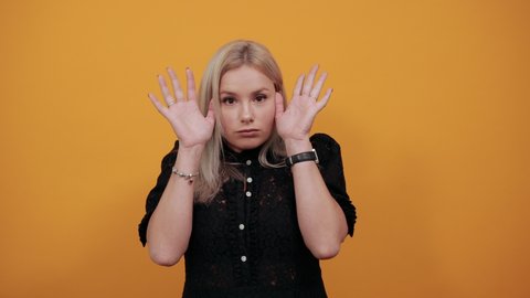 Young blonde girl in black dress on yellow background scared woman is afraid holds her hands to face