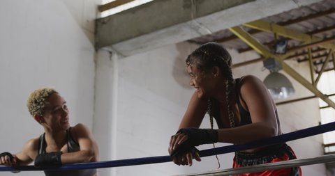 Front view close up of two mixed race female boxers wearing vests, boxing shorts and with hands wrapped, at a boxing gym, leaning on the ropes in a boxing ring, smiling and talking after training,