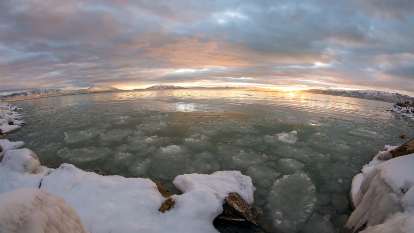 Sunset timelapse over Utah Lake as sky lights up viewing ice in water along snow covered shoreline.