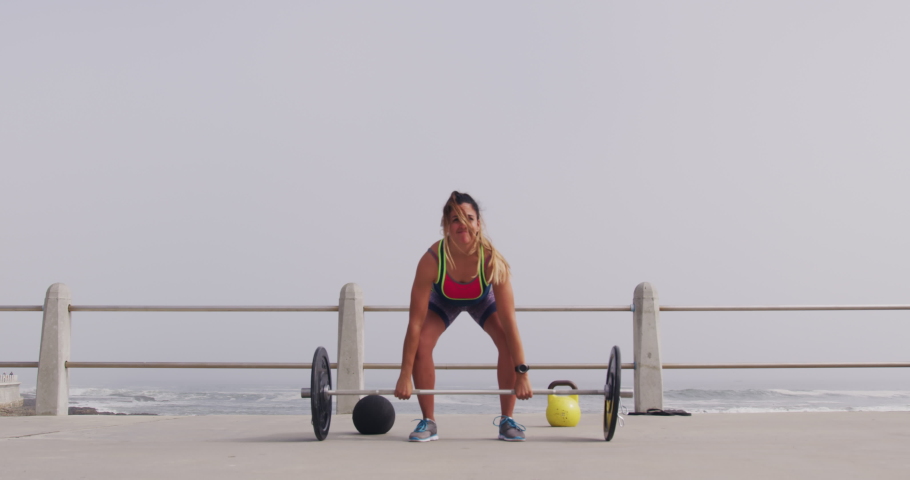 Front view of a young Caucasian woman wearing sports clothes lifting weights during a workout on a promenade, slow motion 