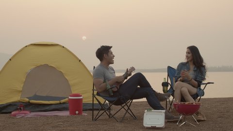 Young Beautiful Asian woman and her boyfriend drinking beer, playing guitar after set up outdoor tent and chair for camping with lake background during sunset in summer season.Sitting together.4k UHD.