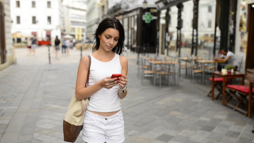 Teen black haired woman in white clothing walking through the calm street, looking around and using smart phone navigation in historical part of Bratislava city. HD VIDEO