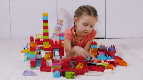 Cute little girl lies on white carpet on floor and playing with colorful lego building blocks. Pretty child having fun playing with plastic constructor. Assembling plastic toy bricks, education toys