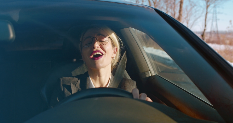 Cheerful and attractive woman in glasses and business style driving a car and singing while listening to the radio on the way to work. | Shutterstock HD Video #1046395750