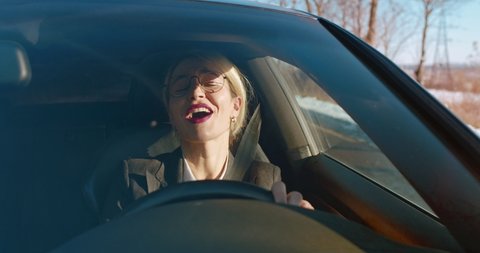 Cheerful and attractive woman in glasses and business style driving a car and singing while listening to the radio on the way to work.