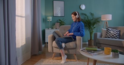 Caucasian young pretty woman in big white headphones sitting in the armchair at the window at home and listening to the music or watching funny video on laptop computer while typing on keyboard. Video de stock