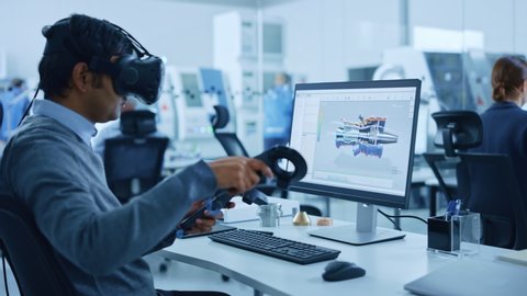 Modern Industrial Factory: Mechanical Engineer Wearing Virtual Reality Headset and Holding Controllers, Uses VR technology for Industrial Design, Development, Prototyping in CAD Software on Computer.