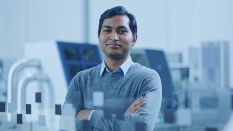 Modern Factory Office: Portrait of Handsome Indian Engineer Crosses Arms and Smiles Charmingly. In the Background High Tech Facility with CNC Machinery, Robot Arm and People Working