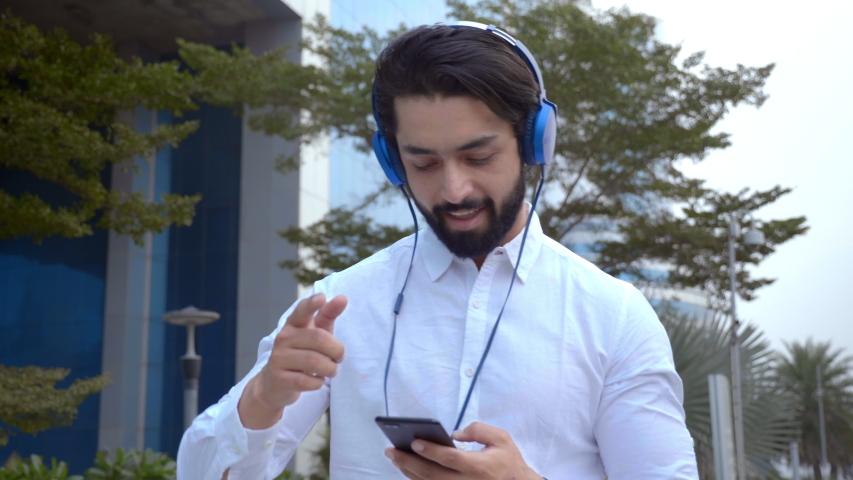 An outgoing casual Indian man wearing headphones smiling and dancing while listening to music. A smiling good looking young male grooving to upbeat music on mobile phone while walking outdoors. Royalty-Free Stock Footage #1046427517