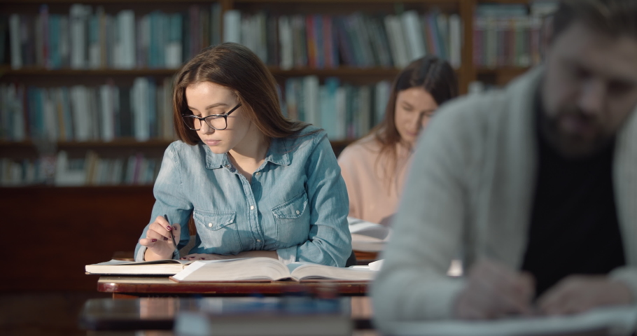 Young pretty female in eyeglasses and blue denim shirt studying in library, education