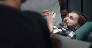 Close up of bearded mature patient lying on grey sofa at psychologist office and sharing his emotions with all pain and frustration. Depressed man visiting skillful doctor during hard time.