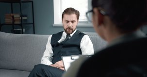 Back view of female psychologist listening carefully emotional bearded patient in formal clothing that sitting on grey couch. Mature man consulting with competent doctor about depression.