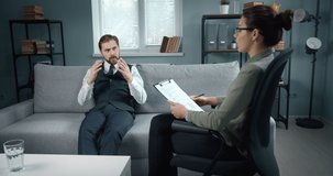 Mature bearded man in formal clothing sitting on grey couch at psychologist's office and gesturing while talking about his problems. Female doctor taking notes and listening carefully.