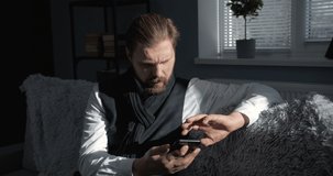 Serious bearded man in white shirt, sleeveless and knitted scarf sitting on sofa at home and using smartphone. Mature male solving important work issues online with modern technologies.