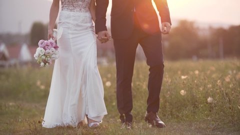 Bride and groom holding hands, walking together with beautiful sunset light background. Slow motion close up shot.